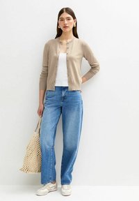 Ljus beige cardigan med rund halsringning, knapp-up front, ihopparad med lösa blå jeans. Modellen håller en vävd väska och bär beige sneakers.