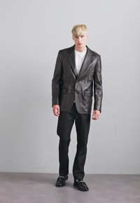 Young man with blonde hair wearing a black leather blazer, white shirt, black pants, and black loafers, standing against a plain gray background.
