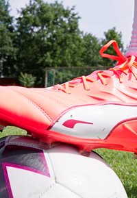 Crampon de football rose vif avec un accent blanc et rouge, upper texturé et lacets visibles, reposant sur un ballon de football noir et blanc à motifs.