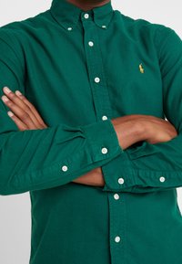 Camisa verde de algodón con botones, que presenta un cuello clásico, botones blancos y un pequeño logo bordado en el pecho izquierdo.