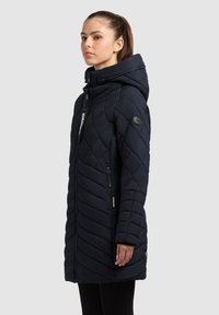 Marineblauw gewatteerde lange jas met een capuchon, voorzien van een ritssluiting aan de voorkant, zijzakken en een logopatch op de mouw. Gladde stofstructuur.