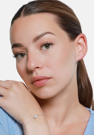 Zilveren armband met een enkele parel, gedragen om de pols. Het model heeft een gemiddelde huidskleur met natuurlijke make-up en lichte blauwe kleding.