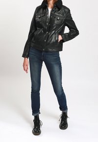 Femme portant une veste en cuir noire avec un col en peau lainée noire, un jean skinny bleu et des bottes noires à lacets, debout avec une main dans la poche.