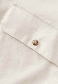 Tissu beige clair avec une texture subtile, comprenant une poche à rabat et un bouton rond marron en accent. Les coutures sont visibles le long des bords.