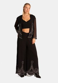 Completo in tessuto nero composto da un top corto e pantaloni a gamba larga, con intricate ricami argentati lungo l'orlo e una giacca lunga trasparente.