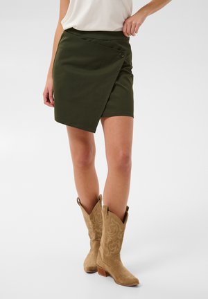 Mujer con falda cruzada asimétrica verde oscuro, top blanco y botas de ante marrón claro estilo western hasta media pantorrilla con bordado.
