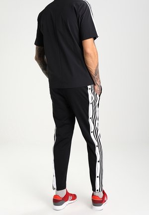 Mand iført sorte træningsbukser og skjorte med hvide striber, røde sneakers, står med hænderne i lommerne, vendt væk.