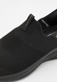 Fekete slip-on cipő texturált kötött anyagból készült, rugalmas talppal, elasztikus oldalsó betétekkel és márkázott húzófüllel rendelkezik.