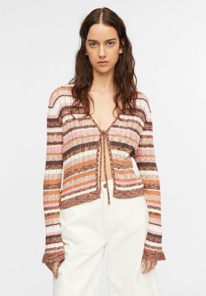 Kobieta o długich ciemnych włosach, nosząca różowo-brązowo-biały prążkowany sweter zapinany z przodu, zestawiony z białymi spodniami z wysokim stanem.