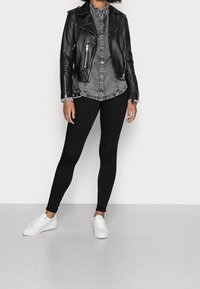 Kvinna som bär en svart läderjacka över en grå knäppskjorta, svarta leggings och vita sneakers, stående mot en enfärgad bakgrund.