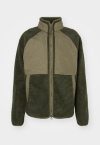 Jachetă din fleece verde cu guler înalt, fermoar complet pe față și design cu panouri contrastante. Textură moale, cu buzunare și manșete ribbate.