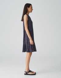 Mouwloze marineblauwe jurk met witte en roze stippen, uitlopende zoom en lichte stof. Gecombineerd met zwarte platte sandalen.