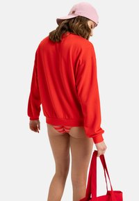 Red oversized sweatshirt with ribbed cuffs, paired with striped bikini bottoms, held with a red handbag, light pink cap worn backward.