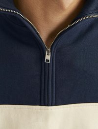 Chaqueta azul marino con panel inferior color crema, que presenta una cremallera metálica y una textura acanalada. Vista en primer plano del cuello y los detalles de la cremallera.