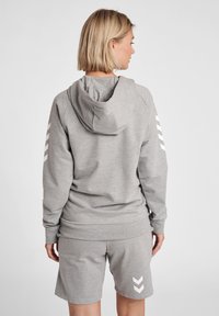 Hoodie cinza com capuz ajustável e detalhes brancos. Combinado com calções cinza com detalhes de chevron brancos nas laterais. Textura de tecido macio.