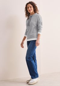 Randig sweatshirt i grått och vitt med huva och dragsko, kombinerad med blå denimjeans och vita sneakers. Lockigt hår synligt.