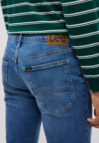 Jeans de denim azul com um corte justo. Apresentam uma etiqueta de couro castanho com o logótipo "Lee", design de cinco bolsos e detalhes de costura visíveis.