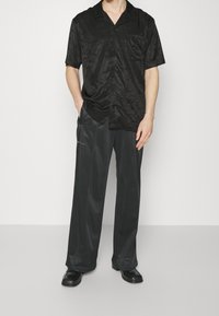 Mustattu lyhythihainen napitettava paita, jossa on hienovarainen tekstuurikuvio, yhdistettynä löysiin, kiiltäviin mustiin housuihin ja mustiin kenkiin.