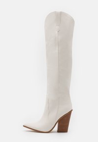Bílá kožená vysoká kozačka sahající až ke kolenu s špičatou špičkou a tvarovaným dřevěným podpatkem. Má švové detaily a strukturovaný horní okraj.