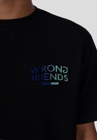 Černé tričko s mnohobarevným vyšitým textem "WRONG FRIENDS DESIGN HOUSE" v různých odstínech modré a zelené. Krátké rukávy.