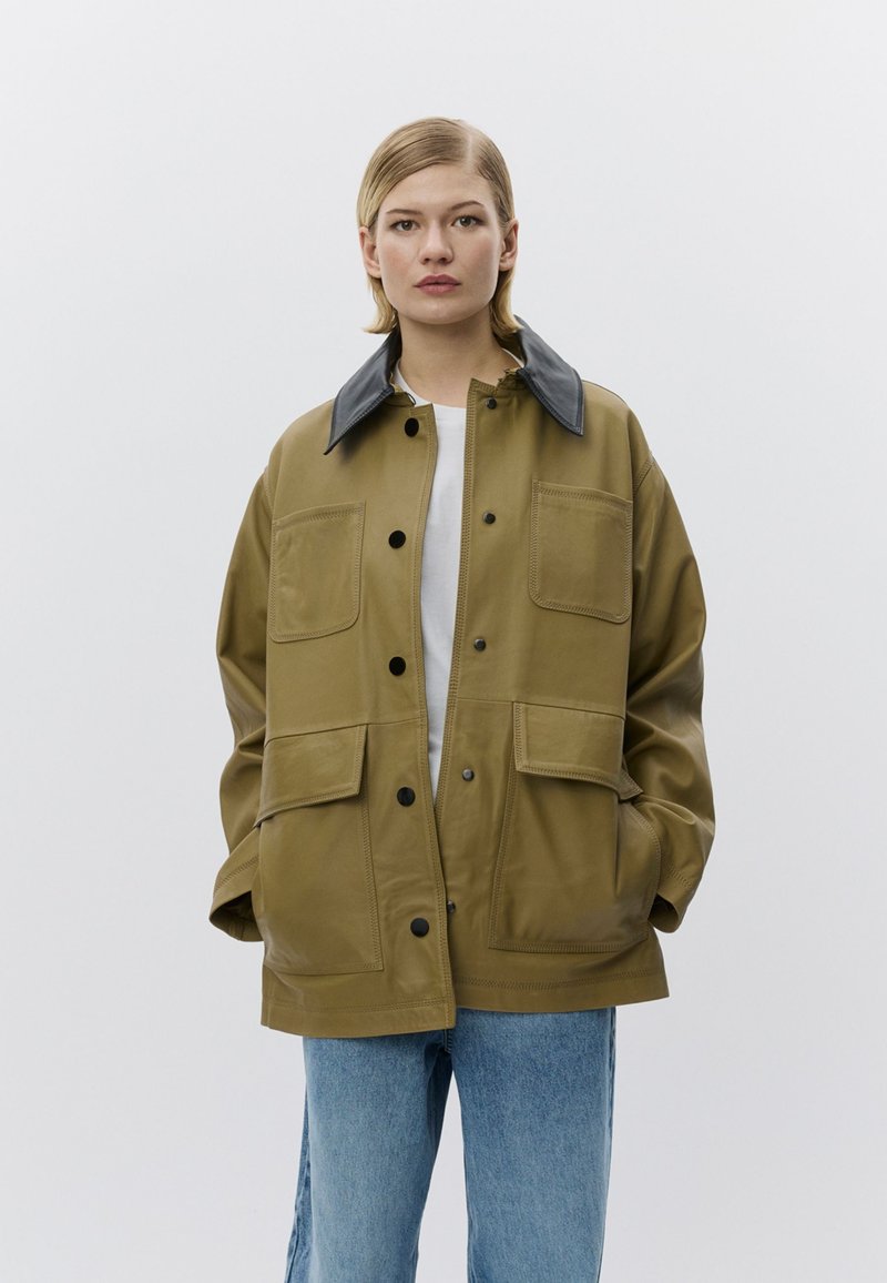 Olivengroene oversized jas met een zwarte leren kraag, voorzien van vier zakken met klep en knoopsluitingen, gemaakt van een glad, duurzaam materiaal.