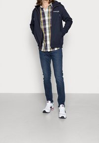 Giacca zip in navy sopra una camicia a quadri con bottoni, abbinata a jeans scuri e sneakers bianche con accenti neri e rossi.