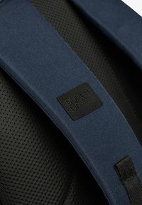 Blå stofrygsækrem med en tekstureret overflade og et sort lædermærke med præget teksten "SUPERDRY." Sort perforeret bagbeklædning synlig.