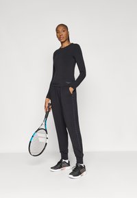 Melnā garām piedurknēm top un sporta bikses, kas izgatavotas no mīksta auduma. Sporta biksēm ir sānu akcenta dizains. Iekļauti melni sporta apavi un tenisa rakete.