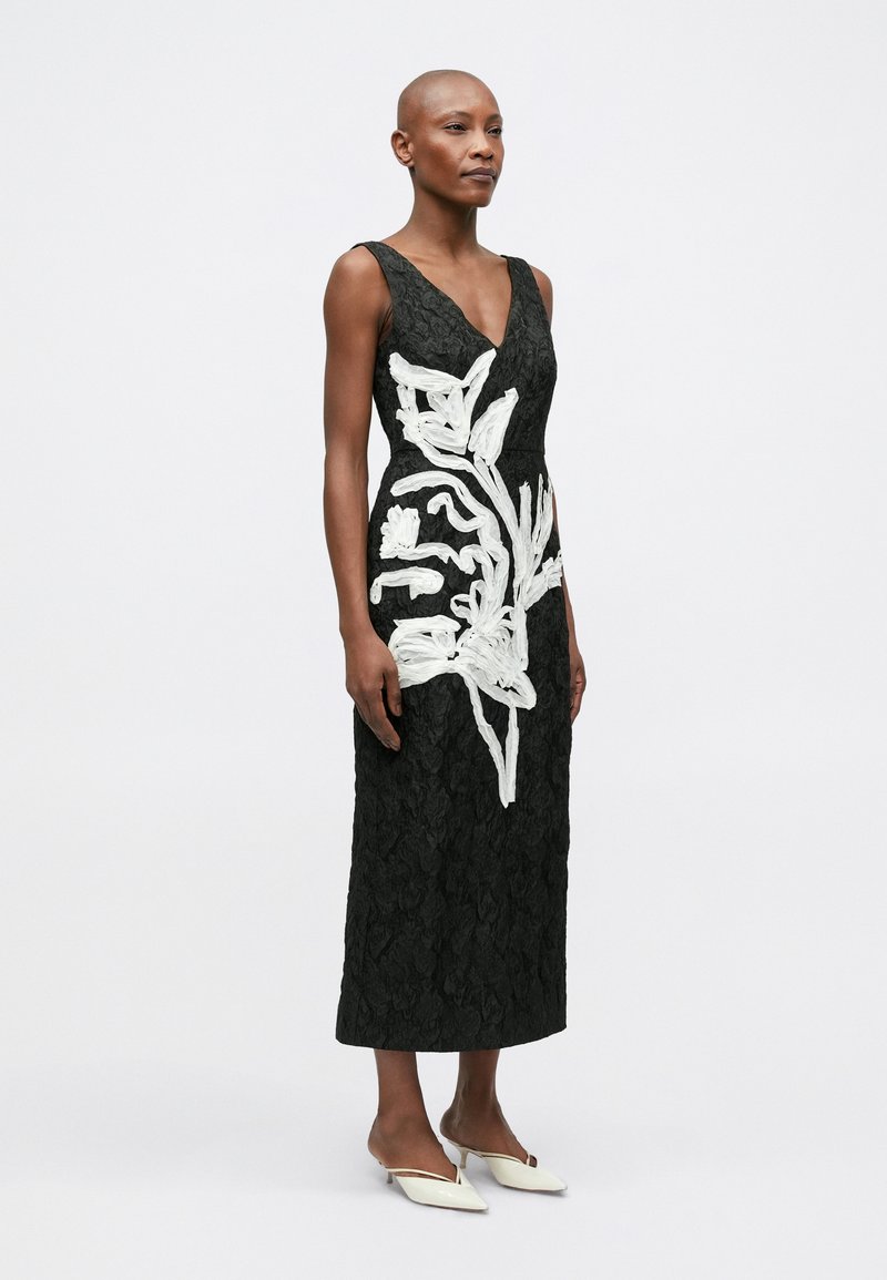 Black lace midi dress featuring white embroidered floral design. V-neckline, fitted silhouette, and textured fabric. Flat white shoes complement.
