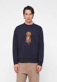 Moški nosi mornarsko modro majico s grafiko rjavega psa, ki nosi tartansko ruto in napis "Hackett", v kombinaciji s bež hlačami.