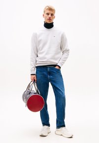 Joven con sudadera gris claro y vaqueros azules sosteniendo una bolsa de deporte azul marino y roja, de pie contra un fondo blanco.