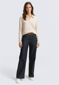 Bež polo majica s dugim rukavima uparena s tamnoplavim širokim trapericama i bijelim tenisicama; smeđi remen naglašava struk.