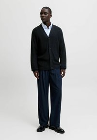 Cardigan en tricot noir avec un col châle, fermeture à boutons et texture côtelée, associé à un pantalon bleu marine à rayures et des chaussures noires.