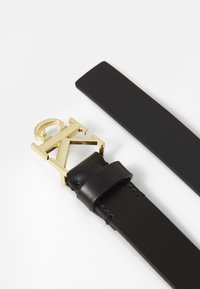 Schwarzer Ledergürtel mit einer goldfarbenen Logo-Schnalle. Der Gürtel ist flach und glatt, mit einem eleganten, minimalistischen Design und ohne Muster.