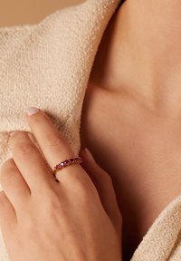 Goldring mit rosa Steinen, der über einen filigranen Band und eine Reihe kleiner runder Edelsteine verfügt. Die Hand ruht auf einem strukturierten, beigen Stoff.