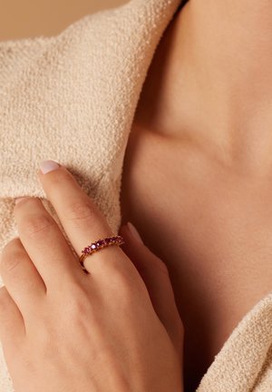 Goldring mit rosa Steinen, der über einen filigranen Band und eine Reihe kleiner runder Edelsteine verfügt. Die Hand ruht auf einem strukturierten, beigen Stoff.