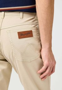 Pantalones Wrangler beige con un parche de cuero que lleva el logo "Wrangler" y costuras decorativas en zigzag en el bolsillo trasero.