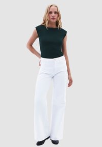 Top verde aderente con maniche corte e scollo alto abbinato a jeans bianchi a gamba larga. Calzature nere con dettagli arrotondati.