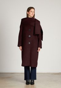 Bordeaux wollen jas met een brede kraag, vier knopen en lange mouwen, gecombineerd met flare jeans en zwarte enkelbotten.