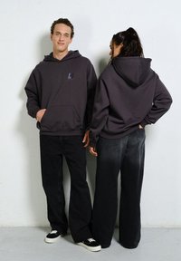 Grafitowa bluza z kapturem z kieszenią z przodu, z małym logo. Połączona z luźnymi czarnymi spodniami jeansowymi. Miękka faktura, codzienny styl.