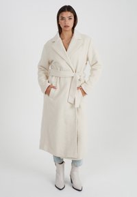 Lungo cappotto avorio realizzato in tessuto morbido e testurizzato. Presenta un design avvolgente, ampi rever e una cintura in tessuto. Due tasche laterali visibili.