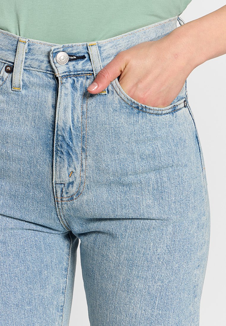 Main dans la poche d'un jean en denim bleu clair taille haute, porté avec un haut vert menthe, montrant les détails du bouton et de la fermeture éclair du jean.