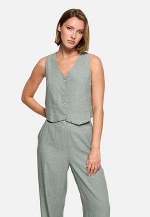 Mujer con chaleco sin mangas color verde claro abotonado y pantalones de pierna ancha a juego de cintura alta, de pie frente a un fondo liso.