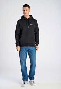 Svart huvtröja med en framfickas kanguroo-liknande design, kontrasterande snören och logotyp. Kombineras med blå jeans och vita sneakers.
