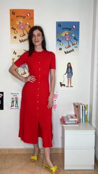 Γυναίκα με κόκκινο φόρεμα με κουμπιά και κίτρινα σανδάλια στέκεται χαμογελώντας δίπλα σε τοίχο με πολύχρωμες αφίσες και βιβλία πάνω σε λευκή ντουλάπα.