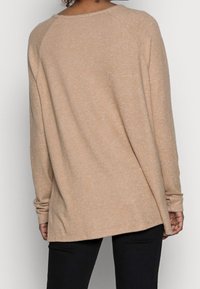 Persona che indossa un maglione beige a maniche lunghe e larghe, con pantaloni neri, vista da dietro su uno sfondo bianco uniforme.