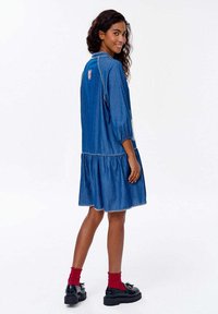 Džínsové šaty s volánovým lemom, 3/4 rukávmi a kontrastným prešívaním. Obsahujú detail na chrbte a sú kombinované s tmavými topánkami a červenými ponožkami.