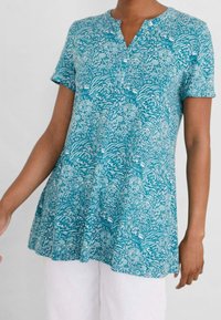 Kvinna i en kortärmad petrolfärgad tunika med intrikata vita blommönster, tillsammans med vita byxor framför en enfärgad bakgrund.