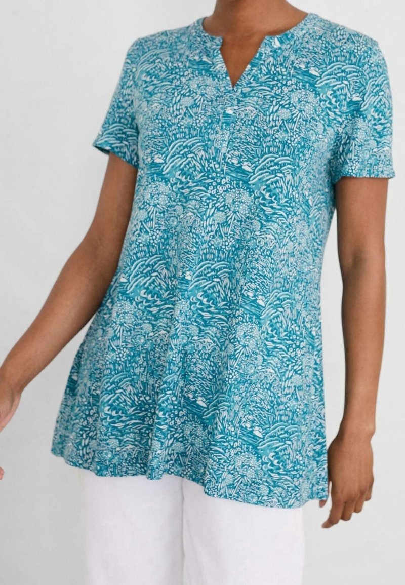 Kvinna i en kortärmad petrolfärgad tunika med intrikata vita blommönster, tillsammans med vita byxor framför en enfärgad bakgrund.