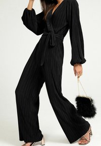Zwarte geplooide jumpsuit met lange mouwen, wikkelontwerp en tailleband. Gecombineerd met metallic hakken en een kleine zwarte nep-bont handtas.